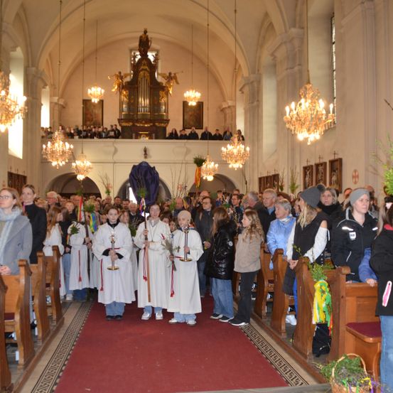 In einer Kirche ist eine große Gruppe von Menschen versammelt. Einige sind in weiße Roben gekleidet und halten Kerzen. Ein roter Teppich verläuft in der Mitte, und Menschen stehen in Bänken auf beiden Seiten.