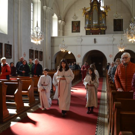 Mehrere Kinder in weißen Gewändern gehen durch den Gang einer Kirche. Ihnen folgen eine Menge Menschen in Winterkleidung. Die Kirche ist mit Kronleuchtern, Gemälden und einer Orgel geschmückt.