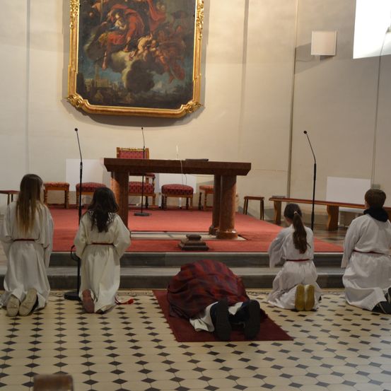 Eine Gruppe von Menschen in weißen Gewändern kniet in einer Kirche, vor ihnen liegt ein Mann auf dem Boden. Ein großes Gemälde ist an der Wand befestigt.