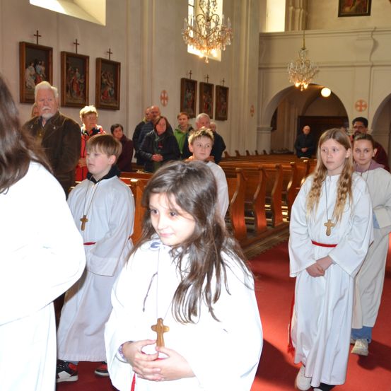 Kinder in weißen Roben mit Kreuzen gehen in einer Kirche. Ein älterer Mann steht hinter ihnen. Mehrere Menschen beobachten von den Kirchenbänken aus.