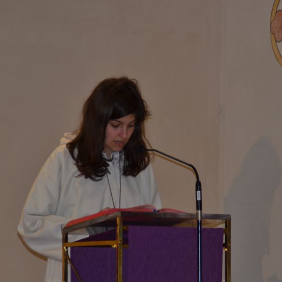 Eine Frau in einer weißen Robe liest von einem Buch an einem Podium mit Mikrofon. Sie sieht einer schlichten weißen Wand zu.