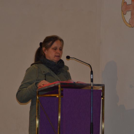 Eine ältere Frau in einer grünen Jacke und einem Schal steht hinter einem Rednerpult mit Mikrofon und liest aus einem Buch.