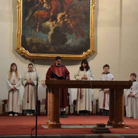 Ein Priester steht vor einem hölzernen Altar mit einem Buch, umgeben von Kindern in weißen Gewändern. Ein großes Gemälde ist an der Wand hinter ihnen angebracht.