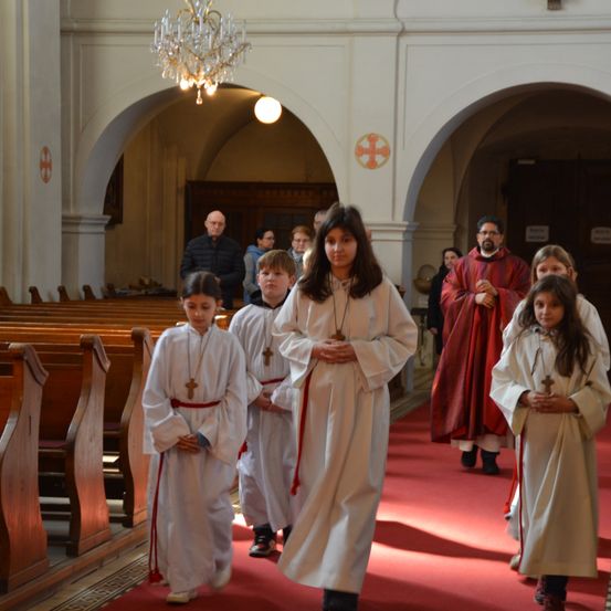 Eine Gruppe von Kindern in weißen Gewändern geht auf einem roten Teppich in einer Kirche. Ein Mann in roten Gewändern beobachtet sie.