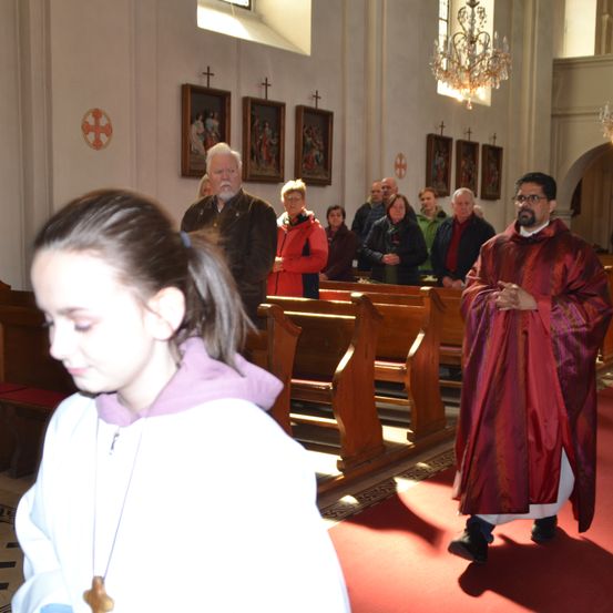 Ein Priester in roten Gewändern steht in einer Kirche und sieht ein junges Mädchen in einem weißen Gewand an. Die Menschen im Hintergrund beten und stehen.