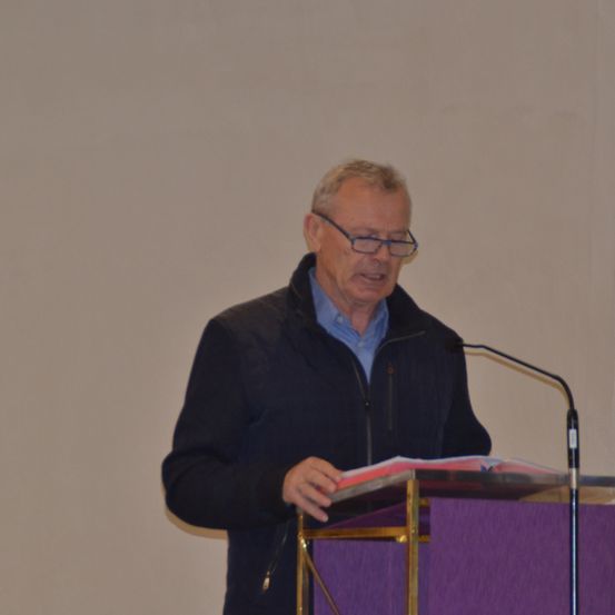 Ein älterer Mann mit Brille steht hinter einem Podium und spricht in ein Mikrofon. Er trägt eine dunkle Jacke über einem hellblauen Hemd.