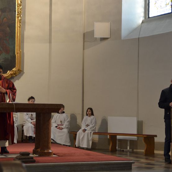 Ein Priester in roten Gewändern steht am Altar in einer Kirche. Einige Leute sitzen auf Bänken, und eine weitere Person steht in der Nähe. Die Kirche hat eine hohe Decke mit einem Gemälde über dem Altar.