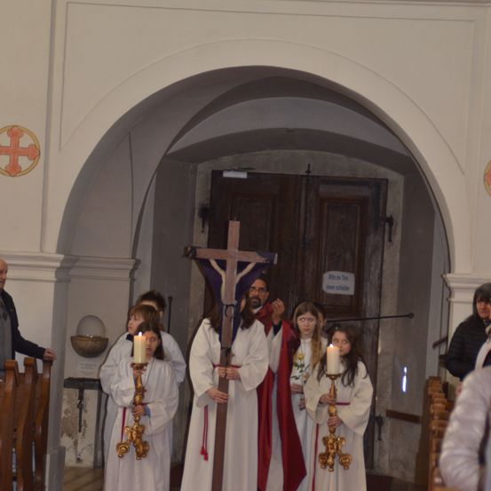 Drei Personen in weißen und roten Gewändern halten brennende Kerzen in einer Kirche in der Nähe einer Holztür mit einem Schild. Ein Bogen und ein Kreuz sind an den Wänden angebracht.