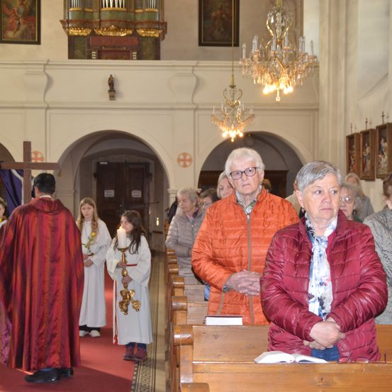 Eine ältere Frau betet in einer Kirche. Ein Priester und junge Kinder halten Kerzen. Die Kirche hat eine hohe Decke mit Kronleuchtern.