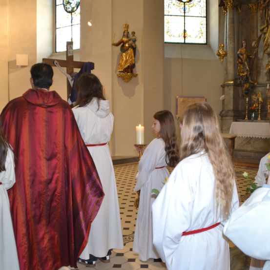 Eine Gruppe von Menschen in weißen Gewändern steht in einer Kirche, angeführt von einem Priester in einem roten Gewand. Ein Kruzifix ist an der Wand hinter ihnen angebracht.
