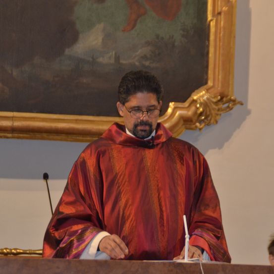 Ein Priester in einer roten Robe und mit einer Brille steht an einem Pult mit einem Mikrofon und liest aus einem Buch. Hinter ihm befindet sich ein großes gerahmtes Gemälde.