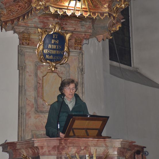 Eine ältere Frau steht an einem Podium in einer Kirche mit einem goldenen dekorativen Kronleuchter über ihr. Der Kronleuchter hat komplizierte Designs und eine blaue Plakette mit goldenen Buchstaben.