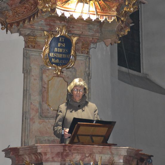 Eine Frau mit Brille steht vor einer dekorativen Plakette über einem goldenen Kronleuchter.