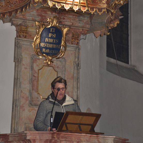 Ein Mann mit Brille steht und schaut nach oben zu einem kunstvollen goldenen Schild mit blauem Hintergrund, das an einer alten Wand befestigt ist.