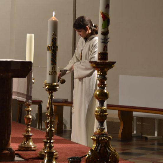Eine Person in einer weißen Robe steht in einer Kirche und hält eine Kerze mit der Aufschrift 2021. Sie zündet eine weitere Kerze mit einem dekorativen Kerzenhalter an.