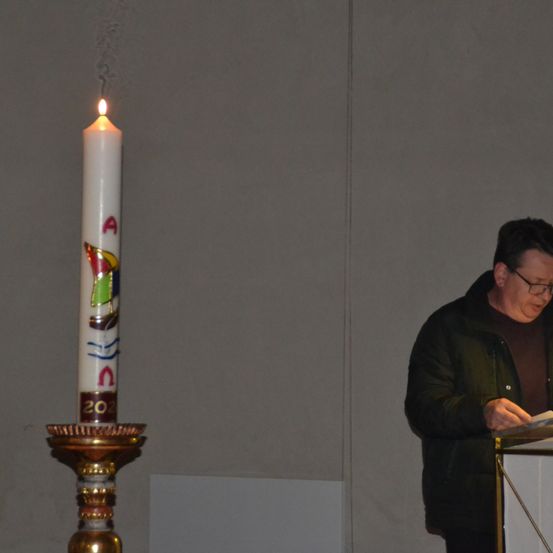 Ein Mann mit Brille steht an einem Rednerpult und liest ein Papier. Eine brennende Kerze mit einem Design steht links von ihm auf einem Ständer.