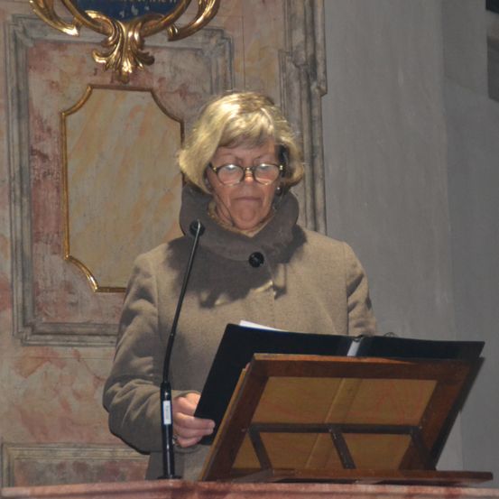 Eine ältere Frau mit Brille und einem grauen Mantel steht an einem Rednerpult und liest aus einem Buch in ein Mikrofon. Der Hintergrund besteht aus einem goldenen Rahmen und Marmorwänden.