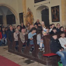 In einer Kirche sitzen Menschen auf Kirchenbänken, einige mit Brille. Eine Statue und ein Gemälde befinden sich an der Wand, und ein roter Teppich liegt auf dem Boden.