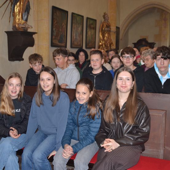Eine Gruppe junger Menschen, sowohl Jungen als auch Mädchen, sitzen in einer Kirche und lächeln für ein Foto. Das Innere der Kirche hat mehrere Gemälde an der Wand und eine Statue auf einem Regal.