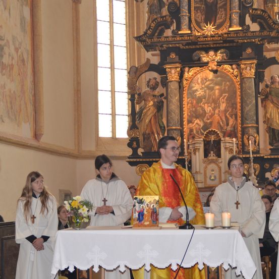 Ein Priester steht vor einem Altar mit zwei Kerzen und zwei jungen Leuten in weißen Roben. Dahinter ein Fenster und ein Gemälde einer religiösen Szene.