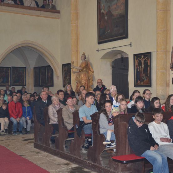 Eine Gruppe von Menschen, einschließlich Kindern, sitzt auf Kirchenbänken in einer Kirche mit Gemälden an der Wand und einer Statue einer Frau, die einen Becher hält.