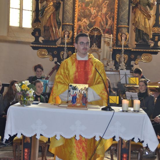 Ein Priester steht an einem Pult, gekleidet in leuchtenden gelben und orangefarbenen Gewändern. Kerzen und Blumen schmücken den Altar vor ihm. Dahinter schmückt eine goldene Wand mit Statuen und kunstvollen Kunstwerken einen heiligen Hintergrund.