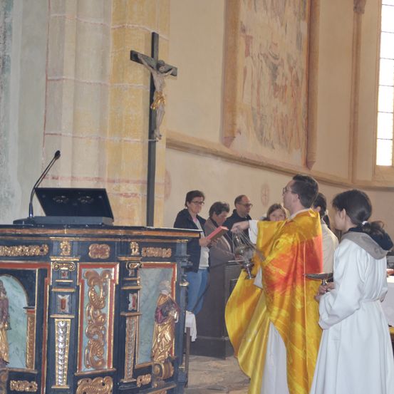Ein Priester in einer gelben und weißen Robe hält ein Weihrauchfass und ein Buch, während eine Frau in einem weißen Gewand einen kleinen Teller hält. Sie stehen in einer Kirche.