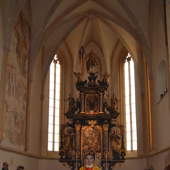 Ein Priester steht am Altar in einer Kirche mit Menschen und Musikern darunter. Der Altar hat kunstvolle Designs und Gemälde.