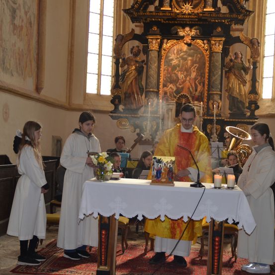 Ein Priester liest an einem Tisch in einer Kirche aus einem Buch, flankiert von zwei Altarservern und zwei Akolythen. Blumen und Kerzen stehen auf dem Tisch, und ein Altarbild befindet sich hinter ihnen.