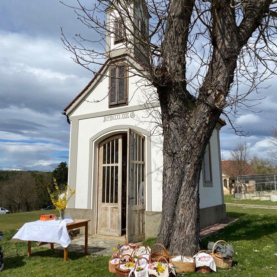 Eine kleine Kapelle mit einer offenen Tür und einem Tisch mit einer Blumenvase davor. Mehrere Körbe sind auf dem Rasen neben dem Baum platziert.