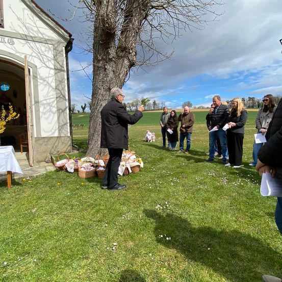 Eine Gruppe von Menschen steht vor einer Kapelle, ein Mann spricht zu ihnen. Körbe und Blumen liegen auf dem Boden. Der Himmel ist teilweise bewölkt.