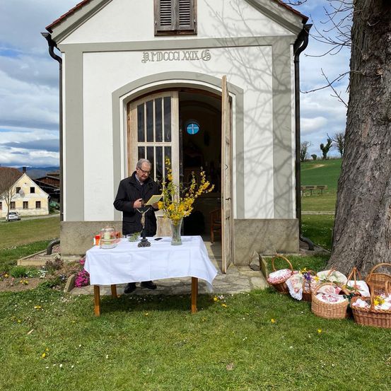 Ein Mann steht vor einer kleinen Kapelle und hält ein Buch. Die Kapelle hat eine offene Tür und ein Kreuzsymbol. Es gibt einen Tisch mit einem weißen Tuch und einer Vase mit gelben Blumen. Körbe mit Blumen sind auf dem Rasen platziert.