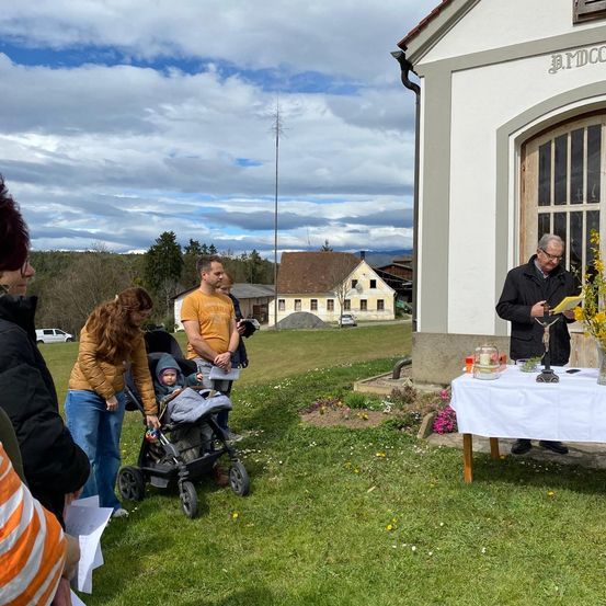 Eine Gruppe von Menschen versammelt sich auf einem Rasen mit einem Baby in einem Kinderwagen und einem Mann, der vor einer Kirche aus einem Buch liest.