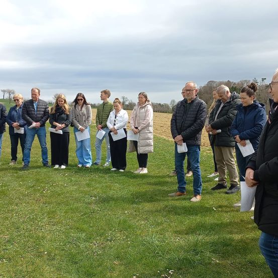 Eine Gruppe von Menschen in Jacken und Jeans steht auf einem Feld und hält Papiere. Sie schauen auf etwas vor sich. Der Himmel ist bewölkt.