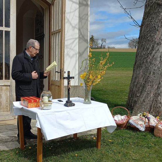 Ein älterer Mann liest ein Buch vor einer Kirche, in der Nähe eines Tisches mit einem Blumenstrauß, einem Kreuz und einem Korb mit Gegenständen. Die Szene spielt sich in einem Grasbereich ab.