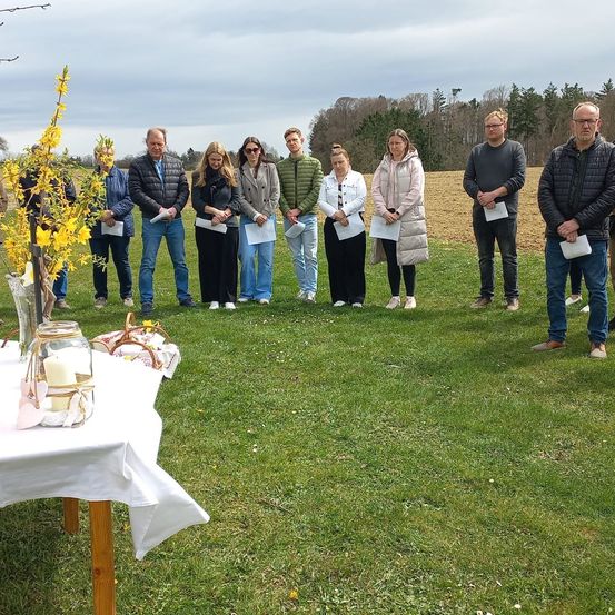 Eine Gruppe von Menschen steht auf einem Feld, hält Papiere in den Händen, mit einem Tisch und einem Korb in der Nähe. Sie sind für kaltes Wetter gekleidet.