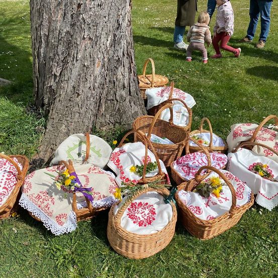 Mit Blumen und Tüchern dekorierte Körbe liegen auf dem Gras, mit Menschen, die um einen Baum im Hintergrund stehen.
