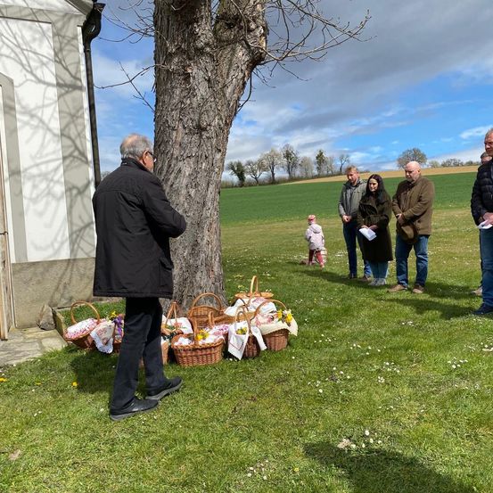 Eine Gruppe von Menschen steht neben einem Baum mit Körben, möglicherweise bei einem religiösen Ereignis. Ein Kind in Pink steht hinter ihnen. Die Szene ist im Freien mit einem grünen Feld und einem bewölkten Himmel im Hintergrund.