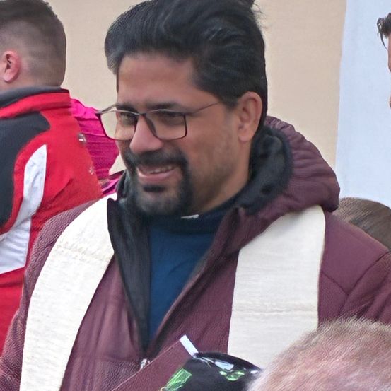 Ein Mann mit Brille und Bart lächelt. Er trägt einen kastanienbraunen Mantel mit einem weißen Schal. Hinter ihm ist teilweise eine andere Person zu sehen, die einen rot-weißen Mantel trägt.