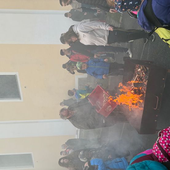 Eine Gruppe von Menschen versammelt sich um ein Feuer, einige tragen Jacken und eine Person scheint das Feuer zu pflegen.