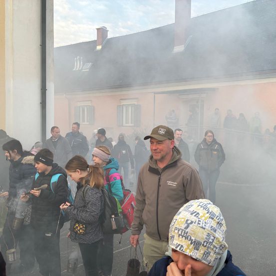 Eine Gruppe von Menschen, darunter Männer, Frauen und Kinder, steht vor einem Gebäude, möglicherweise in einem Dorf, mit Rauch, der aus einem Schornstein aufsteigt. Sie sind warm gekleidet in Winterkleidung.