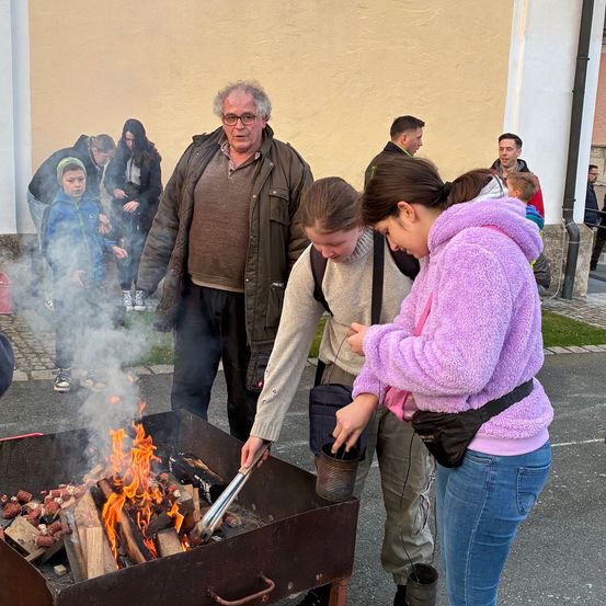 Eine Gruppe von Menschen versammelt sich um einen Feuerkorb und röstet Marshmallows im Freien. Einige tragen Jacken und einer trägt einen Hut.