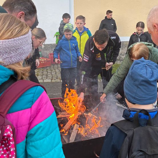 Eine Gruppe von Menschen ist um ein Feuer versammelt, einige tragen Jacken und Turnschuhe, andere haben Mützen. Eine Person hält einen Stock über das Feuer, möglicherweise zum Braten von etwas.