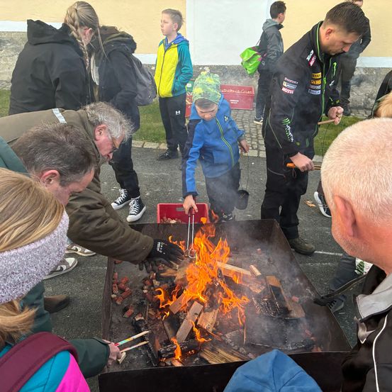Eine Gruppe von Menschen versammelt sich um ein Lagerfeuer und röstet Marshmallows. Ein Kind benutzt Zangen, um das Feuer zu handhaben. Ein Erwachsener hält einen Stock mit einem Marshmallow daran.