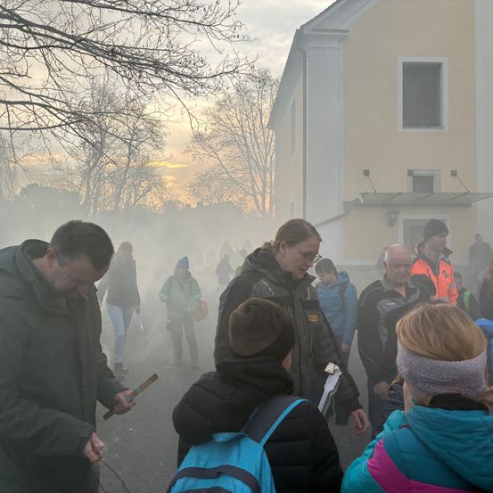 Eine Gruppe von Menschen geht in nebligem Dunst vor einem gelben Gebäude, einige tragen Jacken und Mützen, eine Person hält einen Stock.