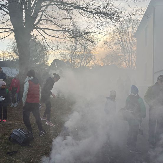 Eine Gruppe von Menschen in Winterkleidung steht in einem nebligen Bereich. Einige beugen sich vor, möglicherweise um ein Feuer zu unterhalten. Rauch entweicht aus dem Boden. Ein Baum und ein Haus sind im Hintergrund zu sehen.