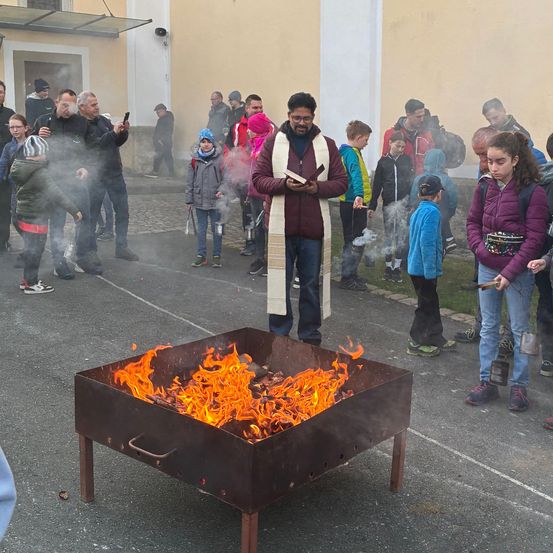 Eine Gruppe von Menschen versammelt sich um ein Feuer im Freien vor einem Gebäude, einige halten Stöcke und Marshmallows.