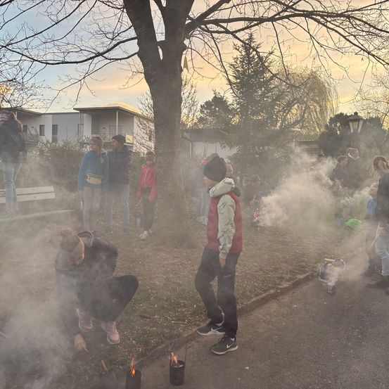 Eine Gruppe von Menschen ist draußen in einem Park, einige knien und entzünden Kerzen vor sich, während andere stehen und zusehen. Bäume, Gebäude und Rauch sind im Hintergrund.