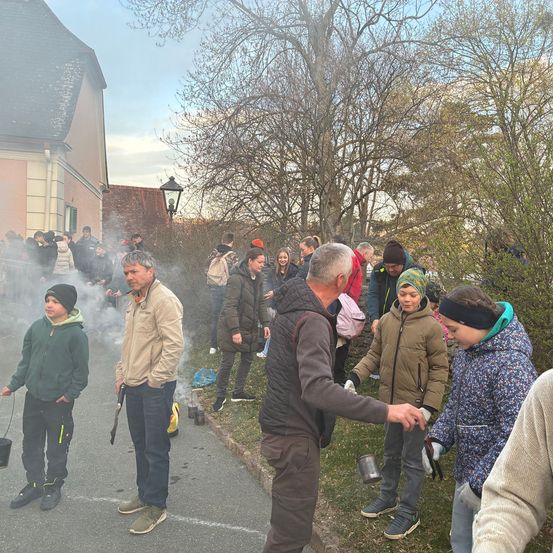 Eine Menschenmenge versammelt sich im Freien, einige halten Tassen und andere stehen bei einem Gebäude. Ein Mann reicht einer Frau eine Tasse. Im Hintergrund befinden sich Rauch und Bäume.