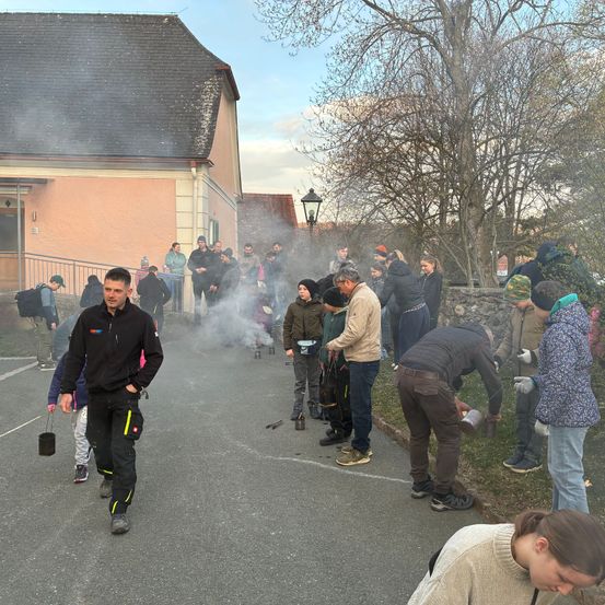 Eine Gruppe von Menschen hat sich draußen versammelt, einige tragen Mützen und Schuhe, ein Mann geht mit einem Becher. Sie sind in der Nähe eines Gebäudes mit einer Lampe und Rauch, der aus dem Boden kommt.